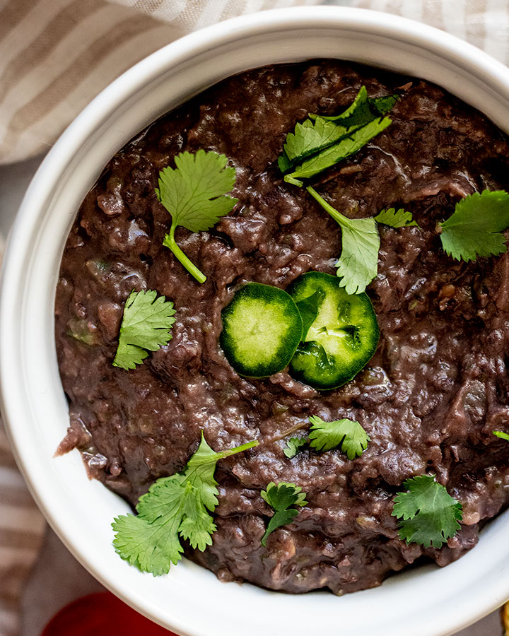 Close up of a bowl of vegan refried beans topped with jalapeno slices and cilantro.