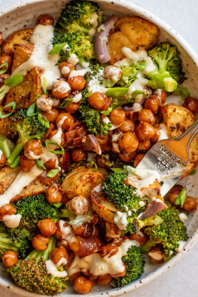 Fork piercing into a roasted potato piece and some broccoli in a bowl of roasted veggies and chickpeas.