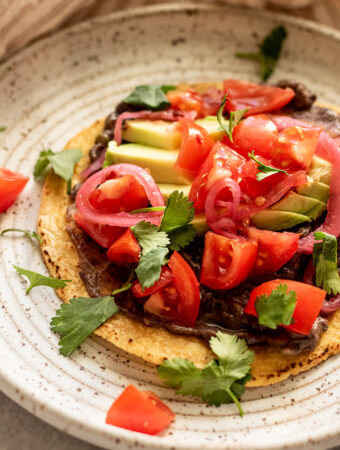 Close up of a layered tostada with refried beans spread on a tortilla with avocado, tomatoes, cilantro and onions on top.