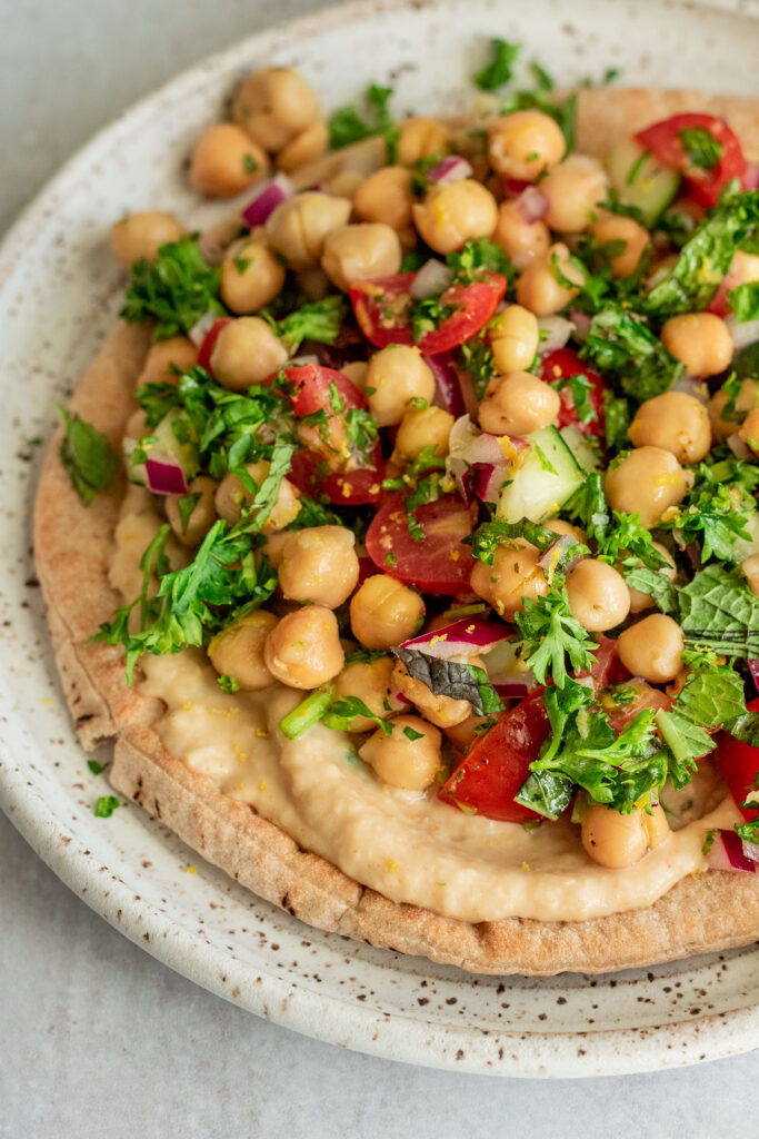 Close up view of a whole wheat pita on a plate smeared with a bean spread and topped with marinated chickpeas and veggies.