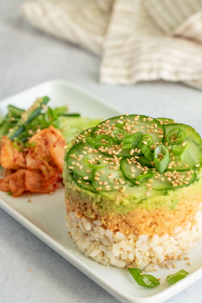 Side view of a rectangular plate with the chickpea stack in front and some fresh cucumbers and kimchi served to the side.