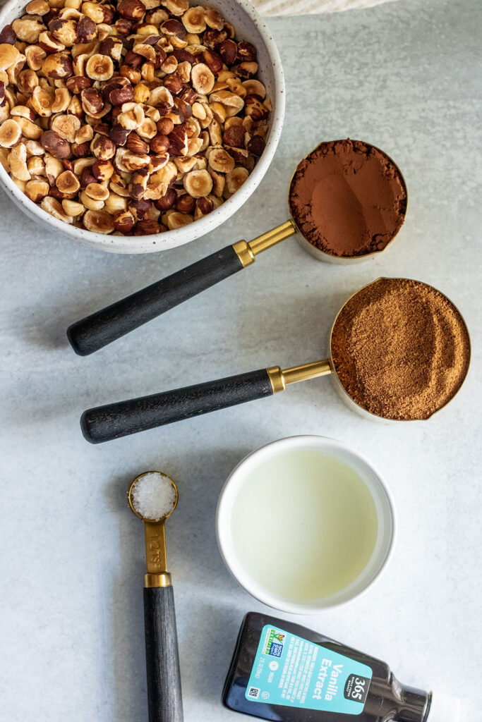 All 6 ingredients placed on a tile surface showing raw hazelnuts, cocoa powder, coconut sugar, oil, salt and vanilla.