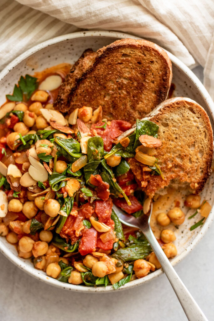 Close up view of a bowl of stewed chickpeas served with crusty bread and topped with parsley and sliced toasted almonds.