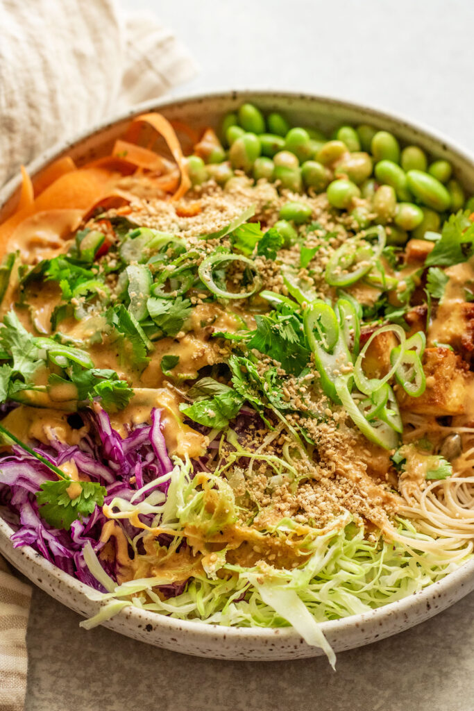 Side view of a bowl of noodles topped with shredded vegetables, edamame, scallions and sesame sauce.
