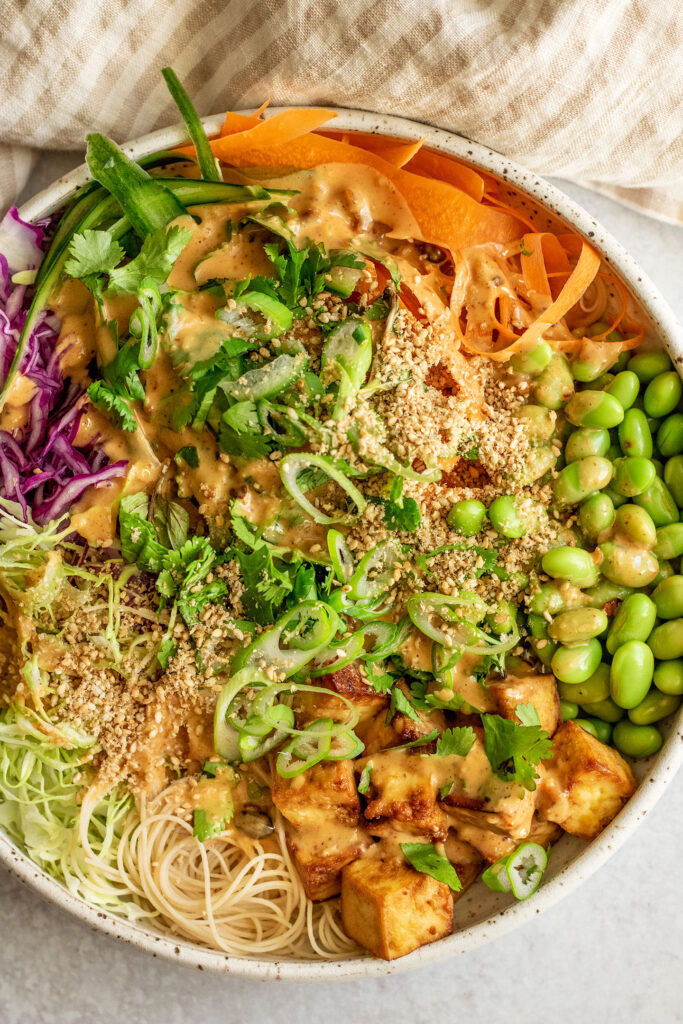 Large bowl of shredded vegetables served with noodles, edamame, tofu and topped with scallions and sesame sauce.