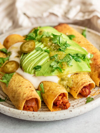 Close up view of 4 flautas on a plate topped with vegan sour cream, avocado slices, cilantro, and pickled jalapeno.