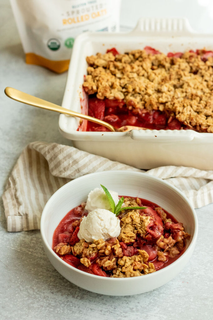 A bowl of strawberry crumble topped with 2 scoops of vanilla ice cream and a piece of mint with the baking dish in the background showing a scoop out of it.