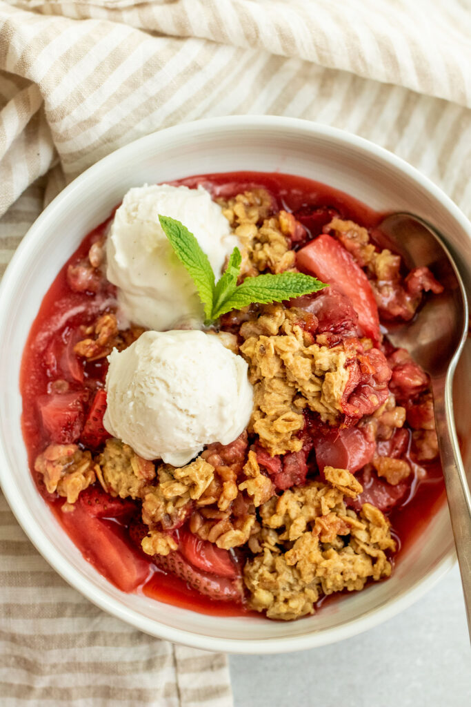 Spoon tucked into the side of a bowl of fruit crumble with two scoops of ice cream on top.