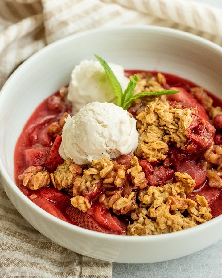 Side view of a bowl of strawberry crumble topped with 2 scoops of vanilla ice cream and mint leaves.
