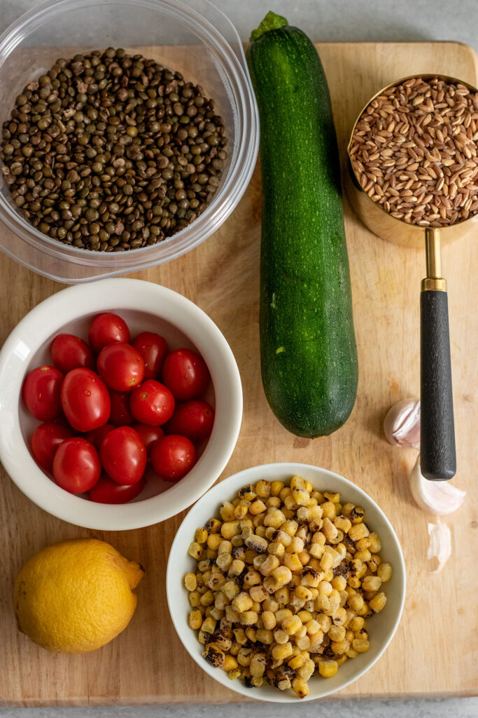 Cutting board with lentils, cherry tomatoes, zucchini, farro, garlic, corn and lemon on top of it.