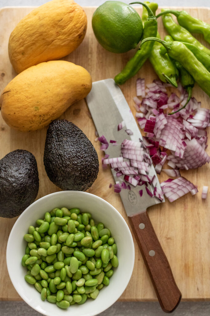 Avocado, mango, edamame, diced onion and shishito peppers on a cutting board with a knife.
