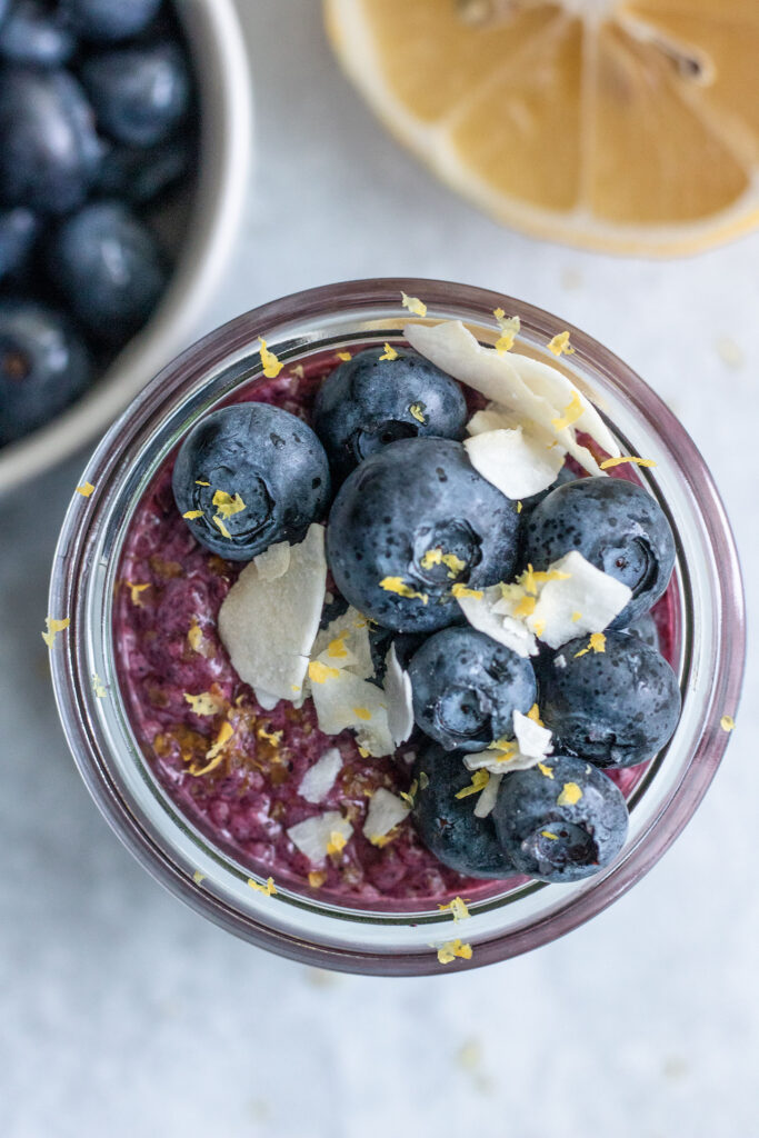 Top down view of a jar of blueberry chia pudding topped with fresh blueberries, lemon zest and coconut flakes.
