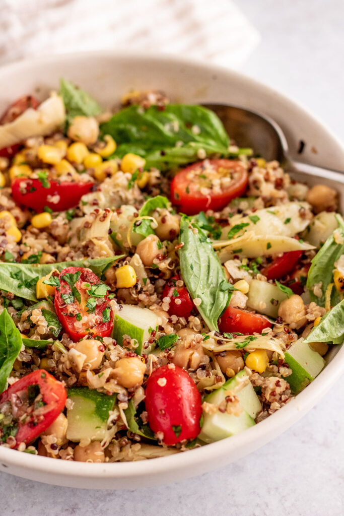 Side view of a bowl of marinated chickpea quinoa tossed with tomatoes, basil, corn and cucumber.