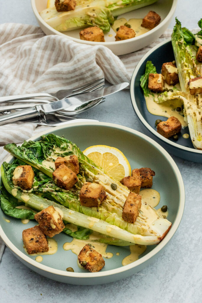Two plates diagonal to each other holding grilled Romaine hearts topped with Caesar dressing and crispy tofu croutons.