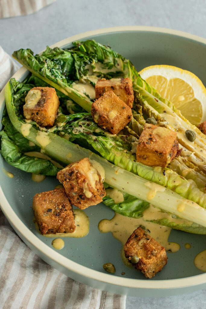 Side view of a plate of grilled lettuce topped with seasoned tofu croutons, a wedge of lemon and extra drizzle of Caesar dressing.