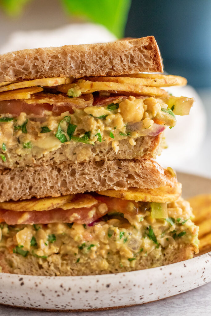 Two halves of a chickpea salad sandwich stacked on each other layered with avocado, chickpea salad, plantains, and tomatoes.