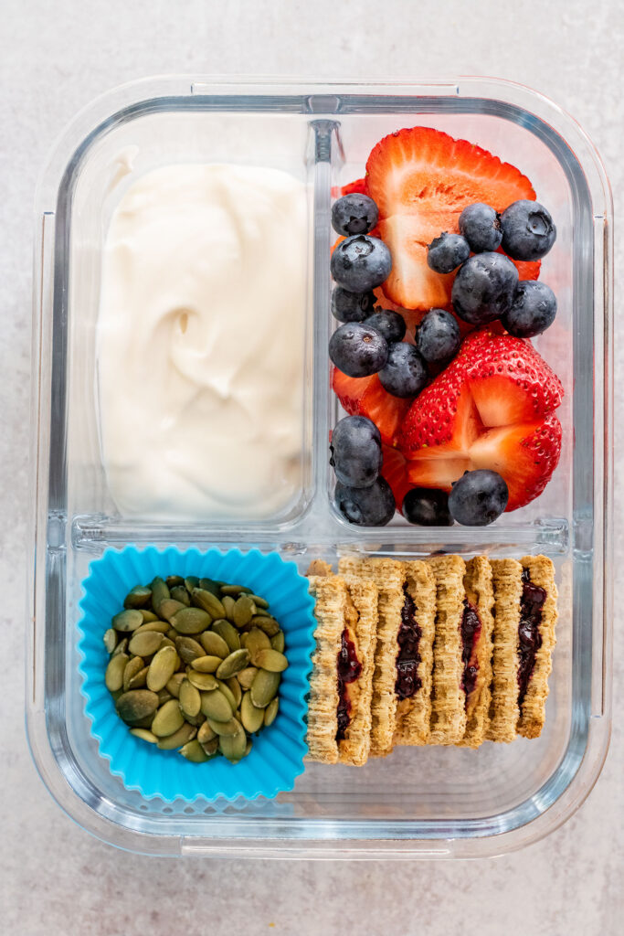 A bento container with one compartment filled with vegan yogurt, another with strawberries and blueberries, another with pepitas and last compartment filled with peanut butter jelly cracker sandwiches.