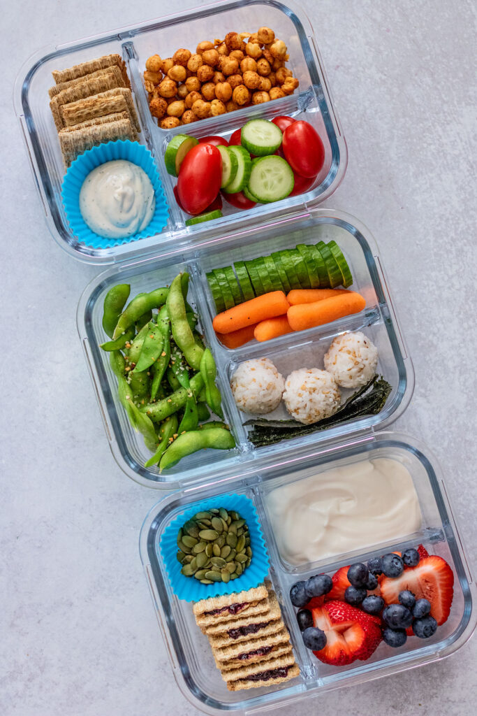 Three glass bento containers filled with a peanut butter cracker sandwich box, a rice ball and veggie box and a roasted chickpeas with veggies and tahini ranch box.