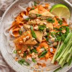 Top down close up shot of a bowl of rice noodles coated in a Red Curry Peanut Sauce and served with pan-fried tofu, sliced cucumbers, scallions and peanuts with a wedge of lime.