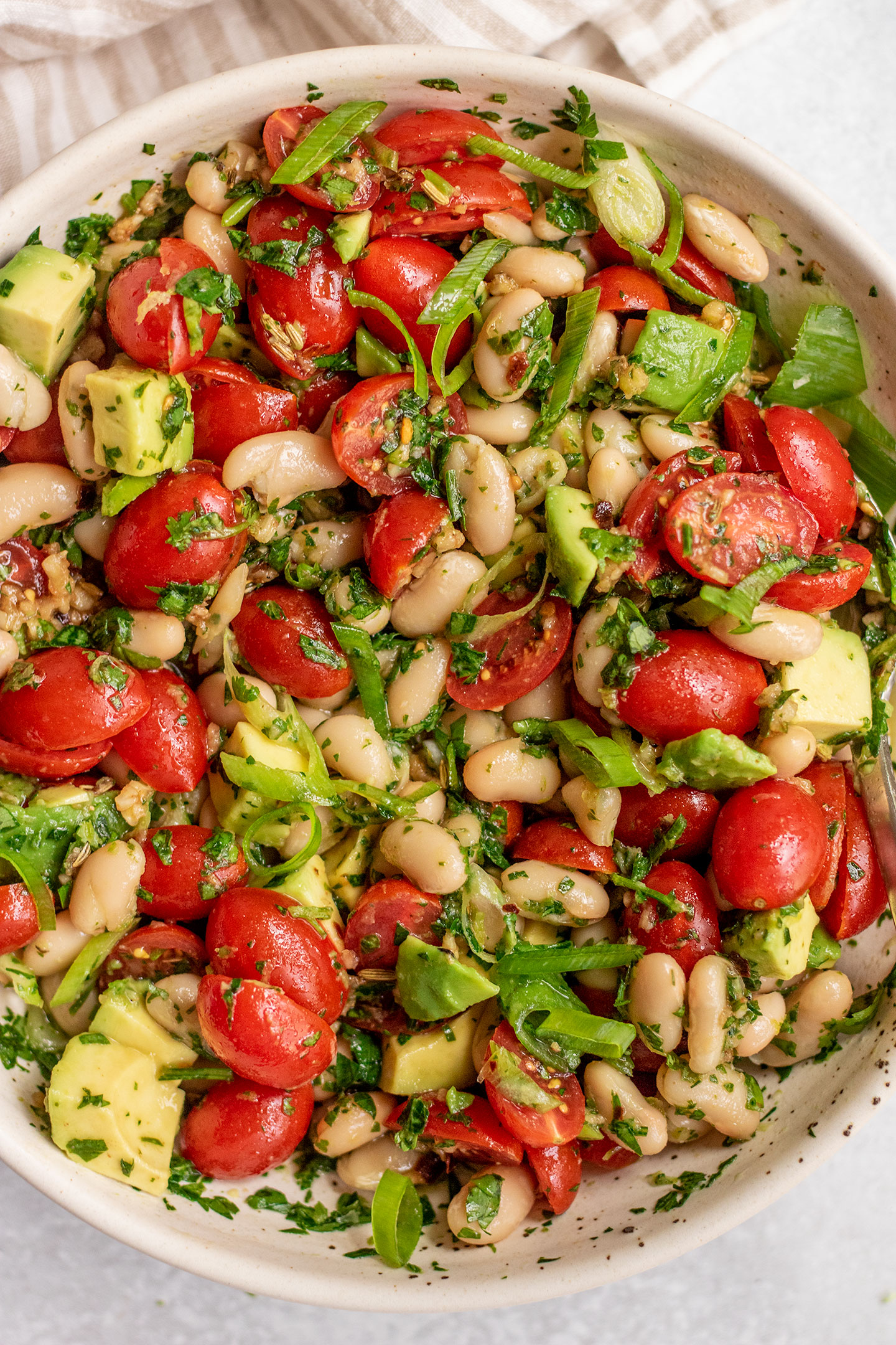 Close up top down view of a bowl of white bean salad mixed with cherry tomatoes, herbs and scallions.