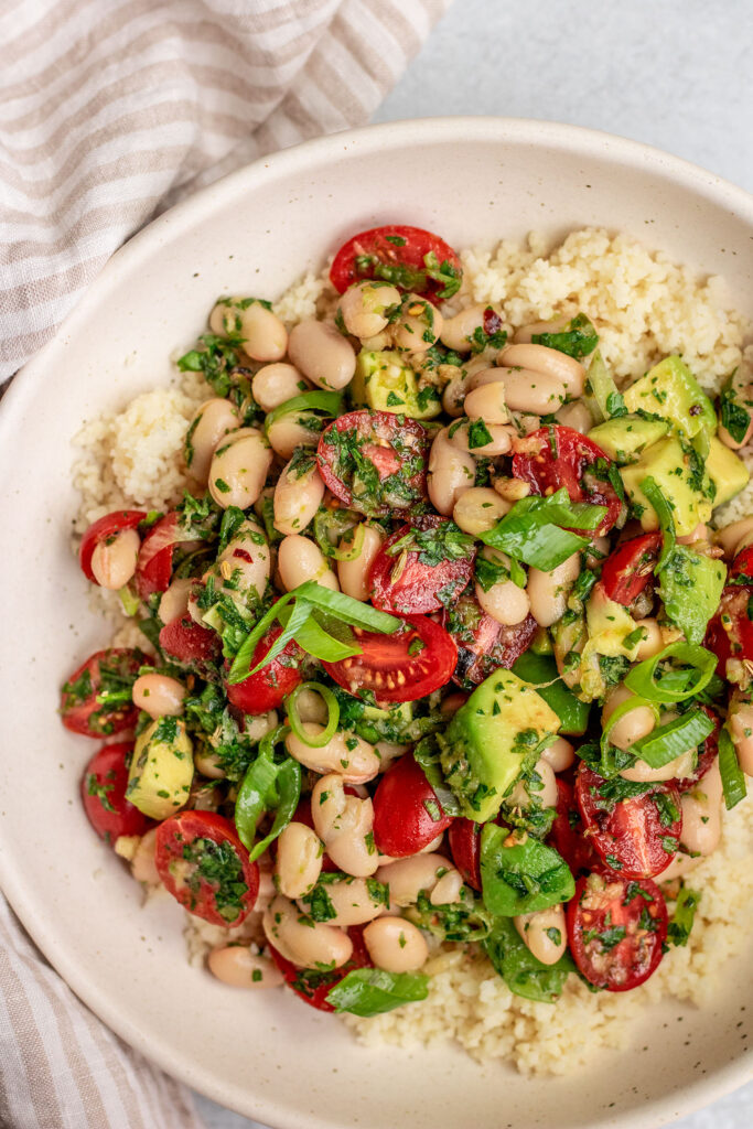 White bean salad topped over couscous in a bowl topped with extra herbs and scallions.