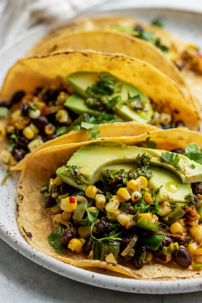Three corn tacos topped with sliced avocado and placed on a white plate sprinkled with cilantro.