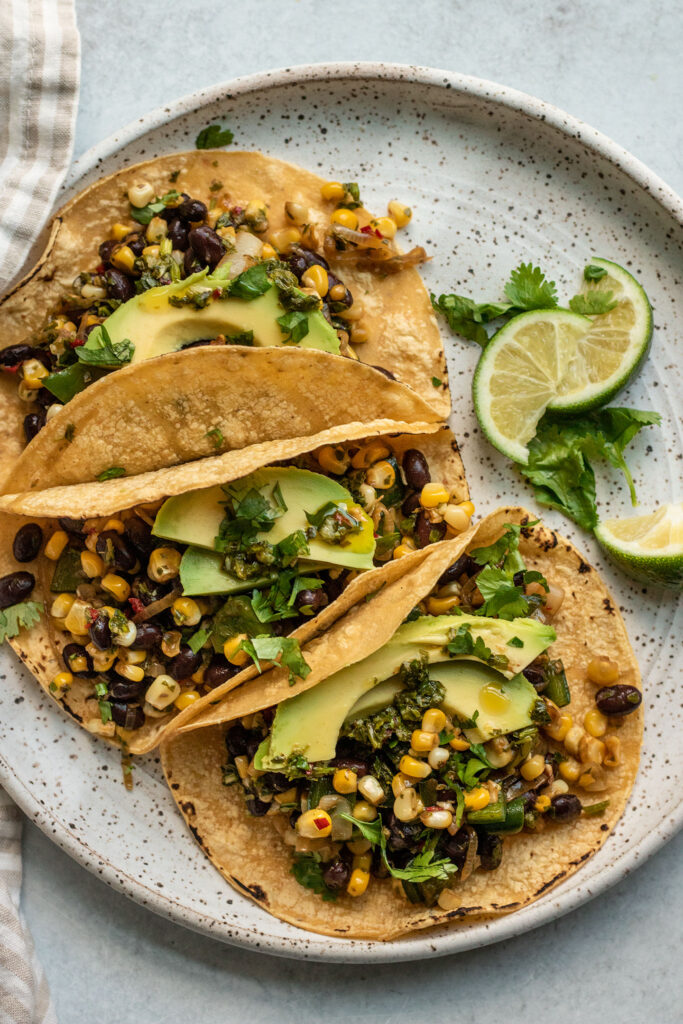Top down view of 3 tacos filled with sauteed corn and peppers mixed with black beans and topped with avocado slices and cilantro.