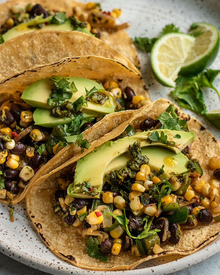 Side view of three corn and black bean filled tacos topped with avocado slices, cilantro and wedges of lime set behind them on the plate.