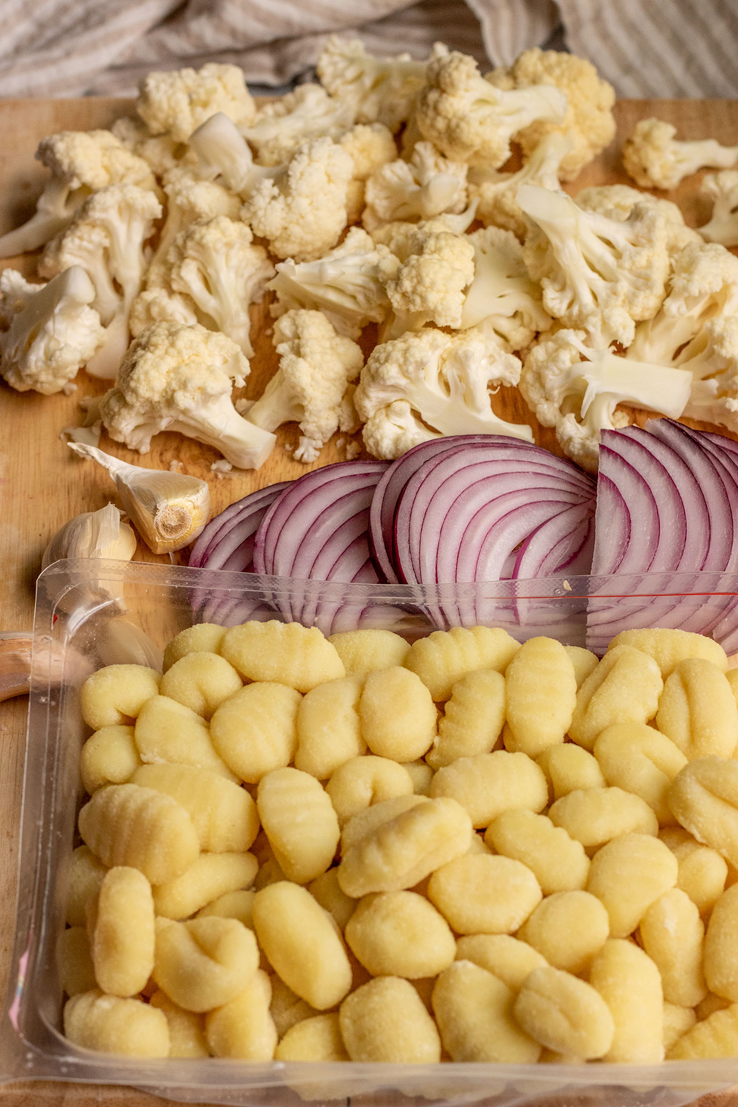 Cutting board topped with sliced onions, cauliflower florets, garlic cloves, and gnocchi.