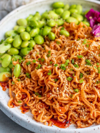 Side view of a bowl of gochujang noodles topped with sesame seeds, chives and served with edamame and pickled cabbage.