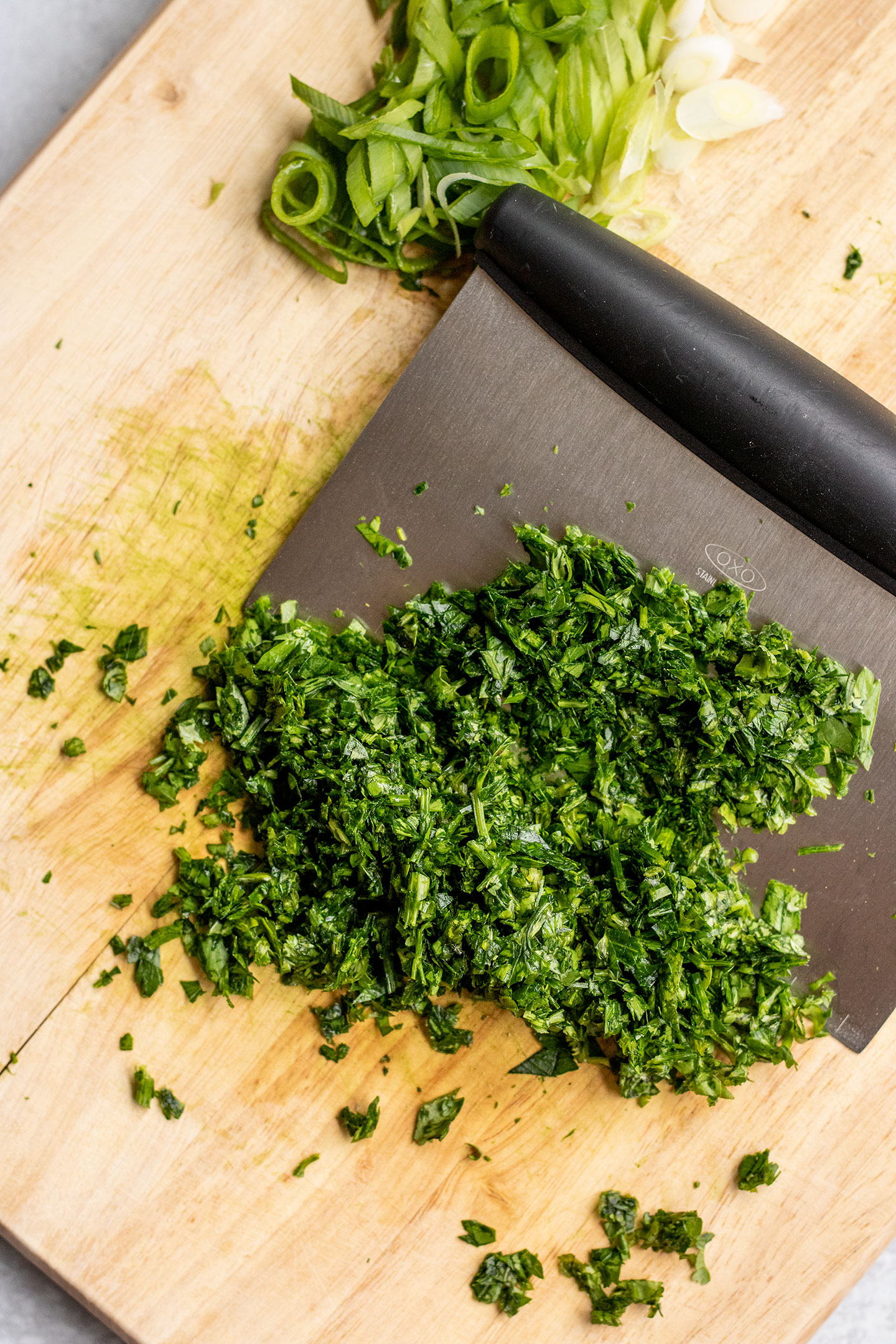 Minced herbs and scallions on a cutting board being scraped with a bench scraper.