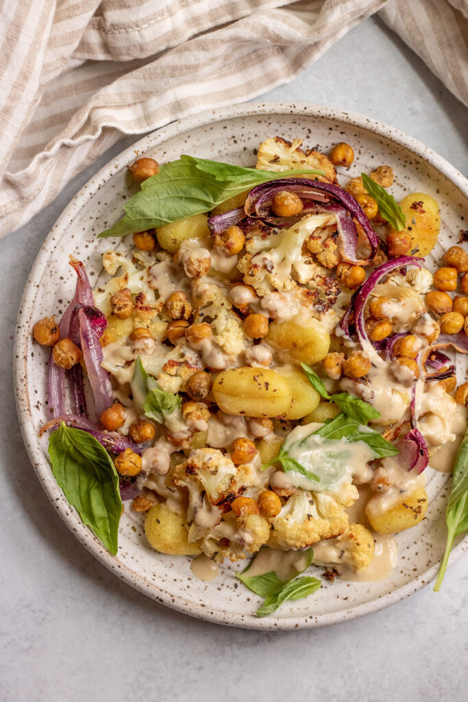 Top down view of a plate of roasted chickpeas, cauliflower, gnocchi and onions topped with garlic tahini sauce.