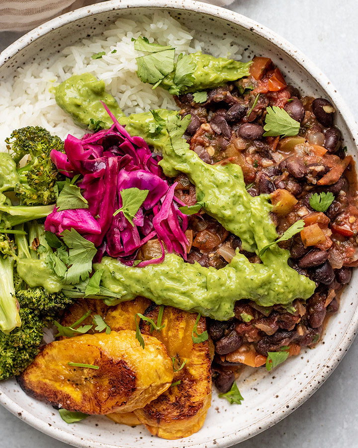 Close up of a plate of rice topped with sofrito black beans, baked plantains, broccoli, pickled cabbage and cilantro lime sauce.