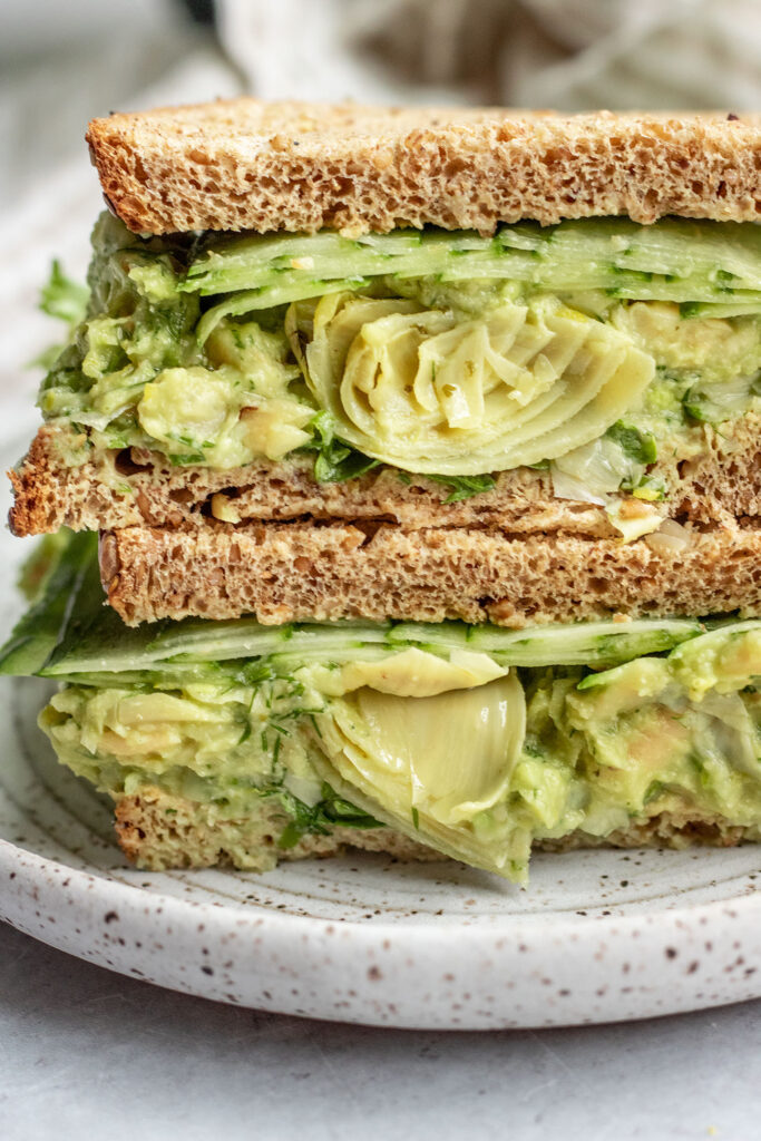 Sandwich cut in half and stacked on top of each other to show it is stuffed with avocado white bean mash, artichoke hearts, greens and cucumbers.