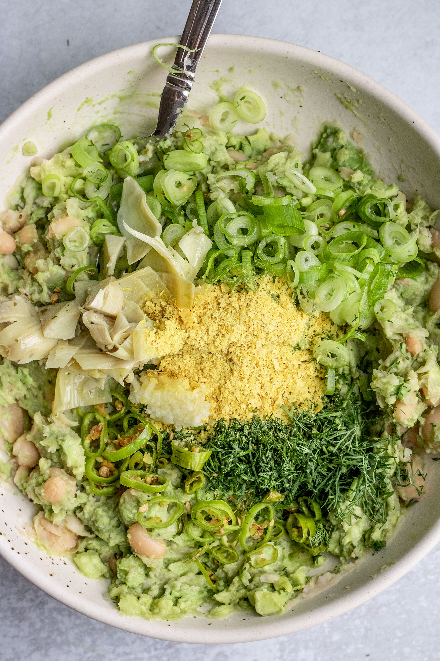 Placing dill, scallions, artichoke hearts, nutritional yeast, peppers, and lemon over top a bowl of mased white beans and avocado.