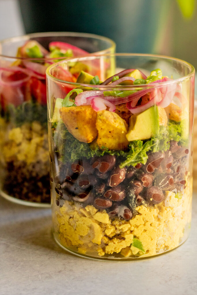 Two breakfast jars, the front facing is filled with scrambled tofu and topped with black beans, potatoes, avocado and pickled onion.