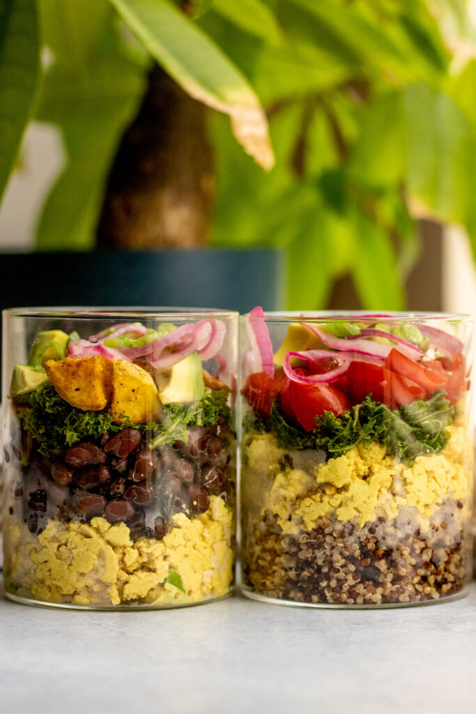 Two different layered savory breakfast jars. The left jar is filled with scrambled tofu, black beans, potatoes, avocado and pickled onion. The jar on the right is filled with quinoa, tofu scramble, massaged kale, cherry tomatoes and red onion.