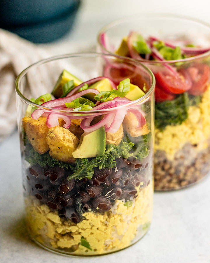 Two jars standing next to each other and loaded with tofu scramble, black beans, kale, potatoes, avocado, and pickled onions.