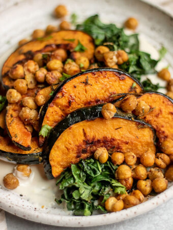 Close up side view of a plate of roasted acorn squash topped with crispy chickpeas and served over a smear of cashew cream and roasted kale.