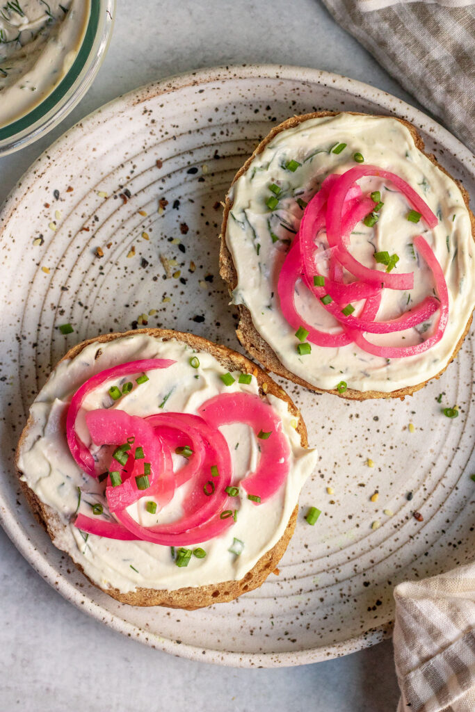 Bagel sliced in half and spread with vegan cream cheese and topped with pickled onions and chives.