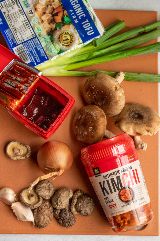 Kimchi, gochujang, mushrooms, scallions, tofu, and onion on a cutting board.