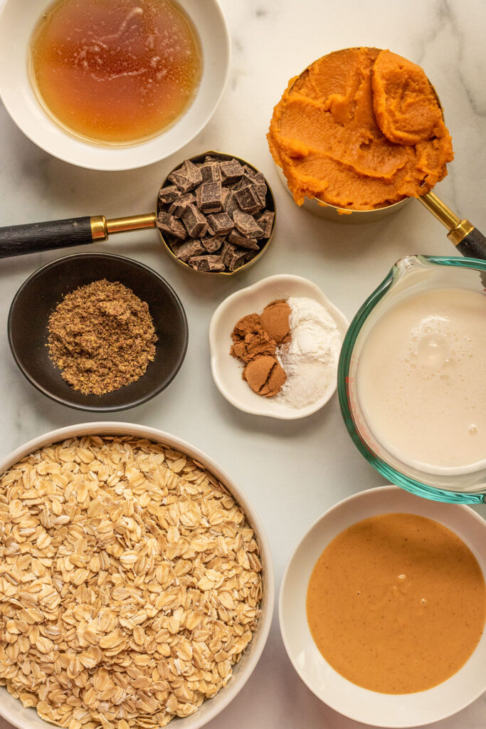 Pumpkin puree, rolled oats, chocolate chips, milk, peanut butter, maple syrup and spices carefully organized on a counter.