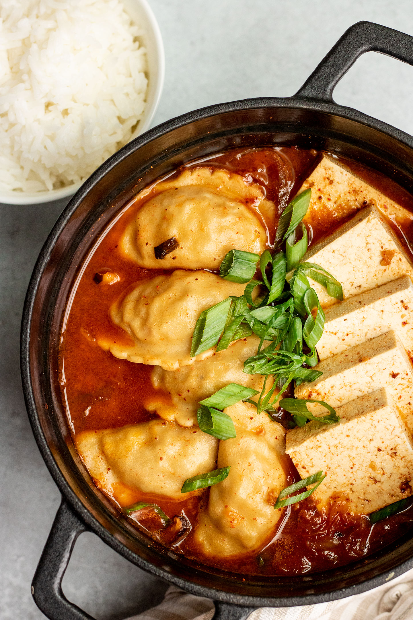 Pot with kimchi stew topped with tofu squares, dumplings and topped with scallions.