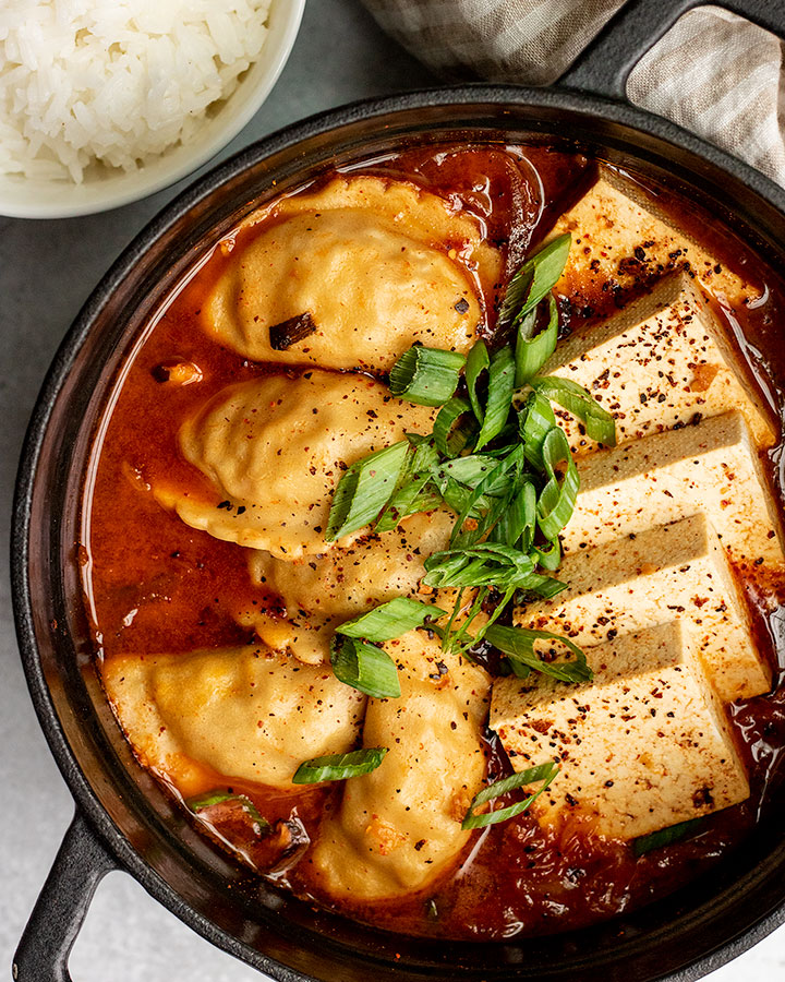 Close up of a pot of kimchi stew topped with tofu squares, vegetable dumplings and garnished with scallions.