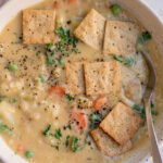 Creamy pot pie soup added to a bowl topped with almond crackers, parsley and black pepper with a spoon tucked in.