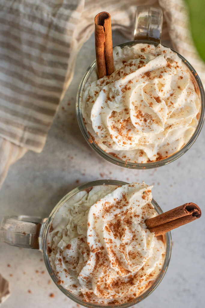 Top down shot of 2 mugs of hot chocolate topped with whipped cream, pumpkin spice and a cinnamon stick for decoration.
