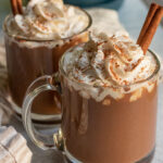 Two mugs of pumpkin spice hot chocolate, in a diagonal of each other topped with whipped cream and pumpkin spice.