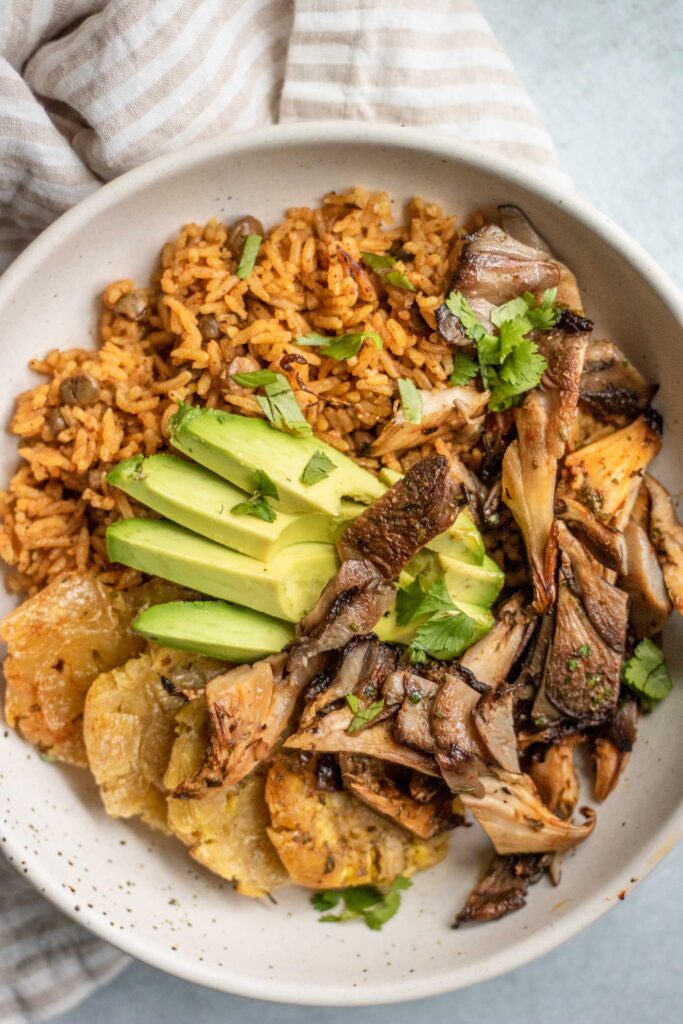 Bowl of rice and beans topped with sliced vegan pernil, plantains, and avocado.