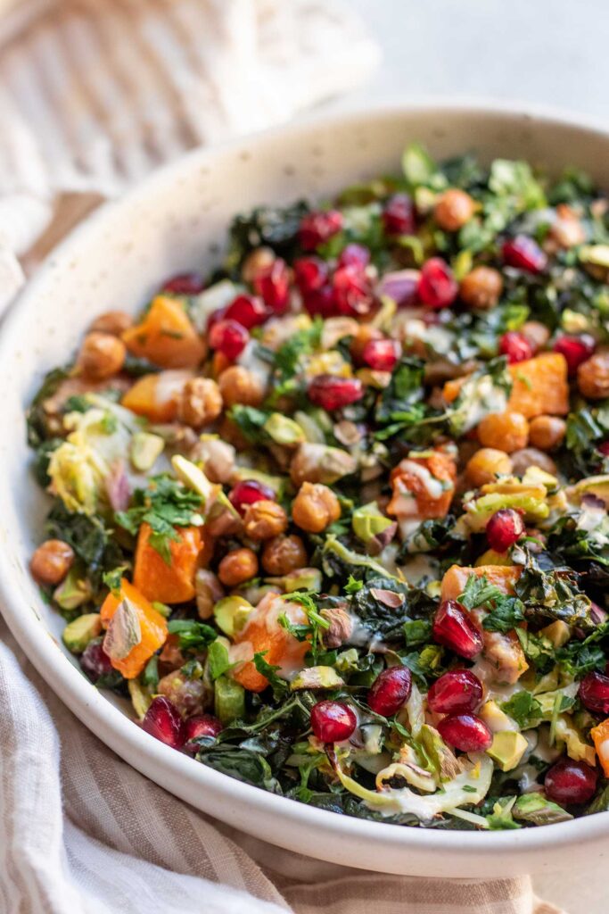 Side view of a kale and brussels sprout salad topped with squash, chickpeas, pomegranate seeds, pistachios and tahini sauce.