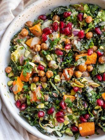 Loaded warm kale salad in a bowl topped with pistachios, pomegranate, parsley and tahini dressing.